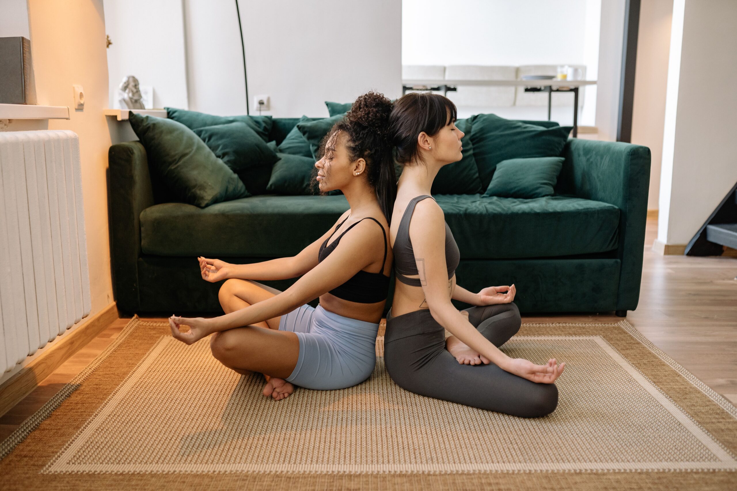 Two women practicing yoga in a peaceful home setting, sitting back-to-back in meditative poses.